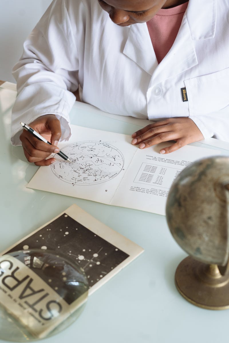 Crop of focused scientist wearing white uniform and modifying constellation map while sitting at table with globe and educational materials in university