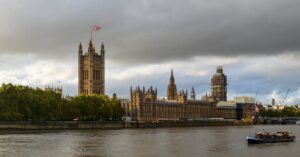 Exterior of Houses of Parliament with flag of England in front of Thames in cloudy day