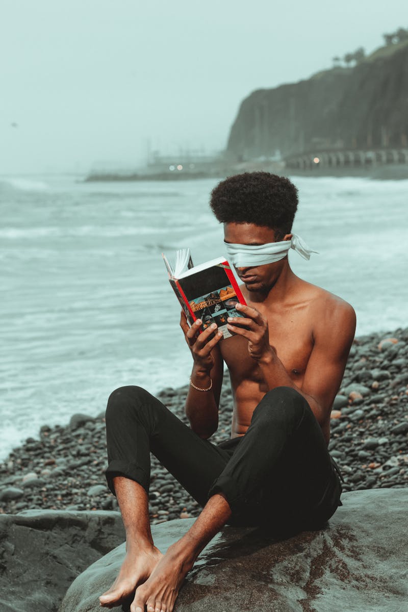 A Blindfolded Man Sitting on the Beach and Holding a Book