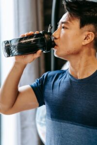 Serious ethnic man in sportswear drinking water from plastic bottle during break in fitness workout in gym